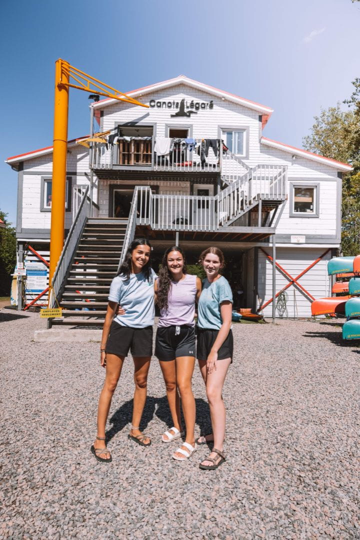 Trois employées Canots Légaré debout et souriantes devant la cabane à canots